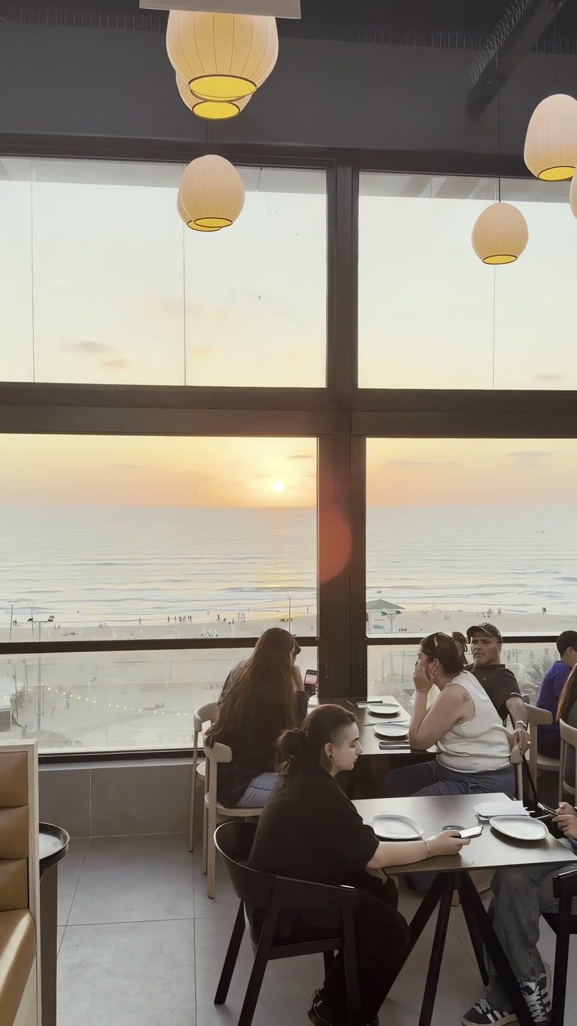 השמש שוקעת אל הים מבעד לחלונות הרופטופ, סועדים ליד החלון